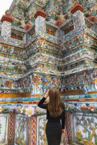 A woman gazes up at Wat Arun, the colorful Temple of Dawn in Bangkok, showcasing its ornate porcelain-covered towers. Must-Visit Temples in Bangkok.