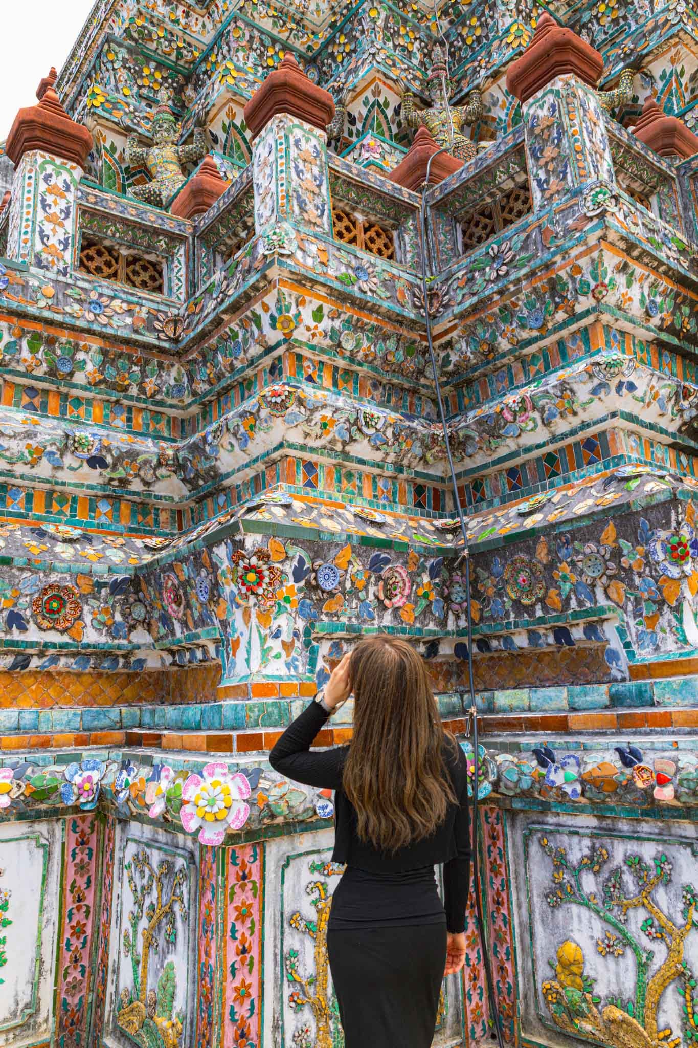 Woman looking up to Wat Arun (Temple of Dawn) in Bangkok, Thailand Full - Yati Travel A woman gazes up at Wat Arun, the colorful Temple of Dawn in Bangkok, showcasing its ornate porcelain-covered towers. Must-Visit Temples in Bangkok.