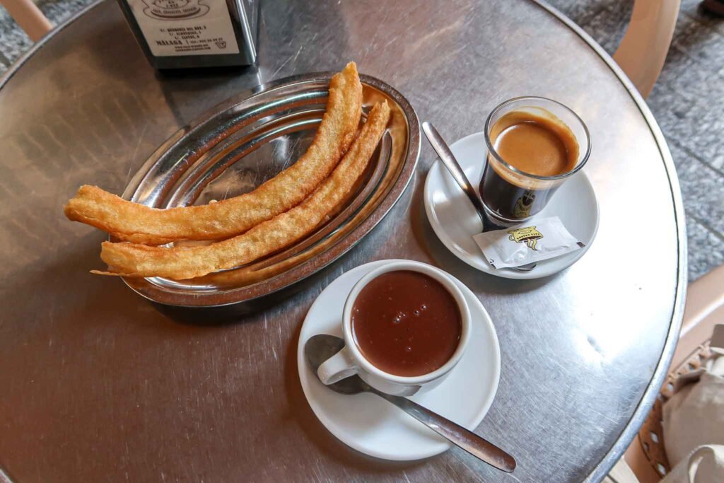 Authentic Churros at Casa Aranda in Málaga, Spain - Yati Travel A table featuring a cup of coffee alongside a delicious churro at Casa Aranda in Málaga, Spain. Best Things to Do in Málaga.