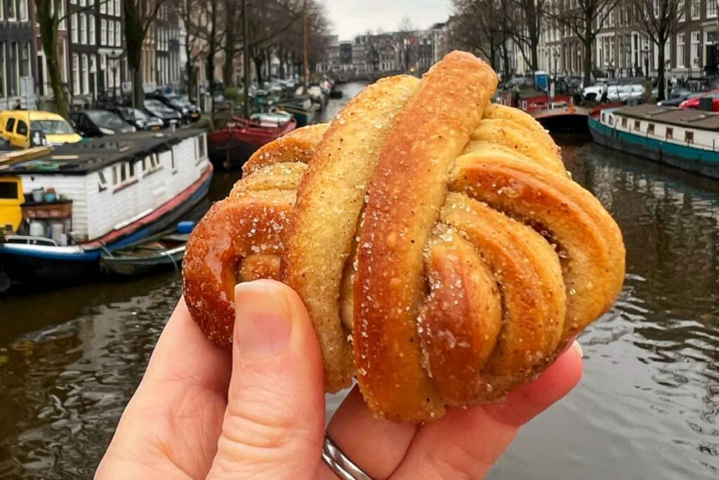 A person enjoys a cinnamon knot pastry by a canal at OSLO Bakery in Amsterdam, surrounded by scenic views. Best Bakeries in Amsterdam.