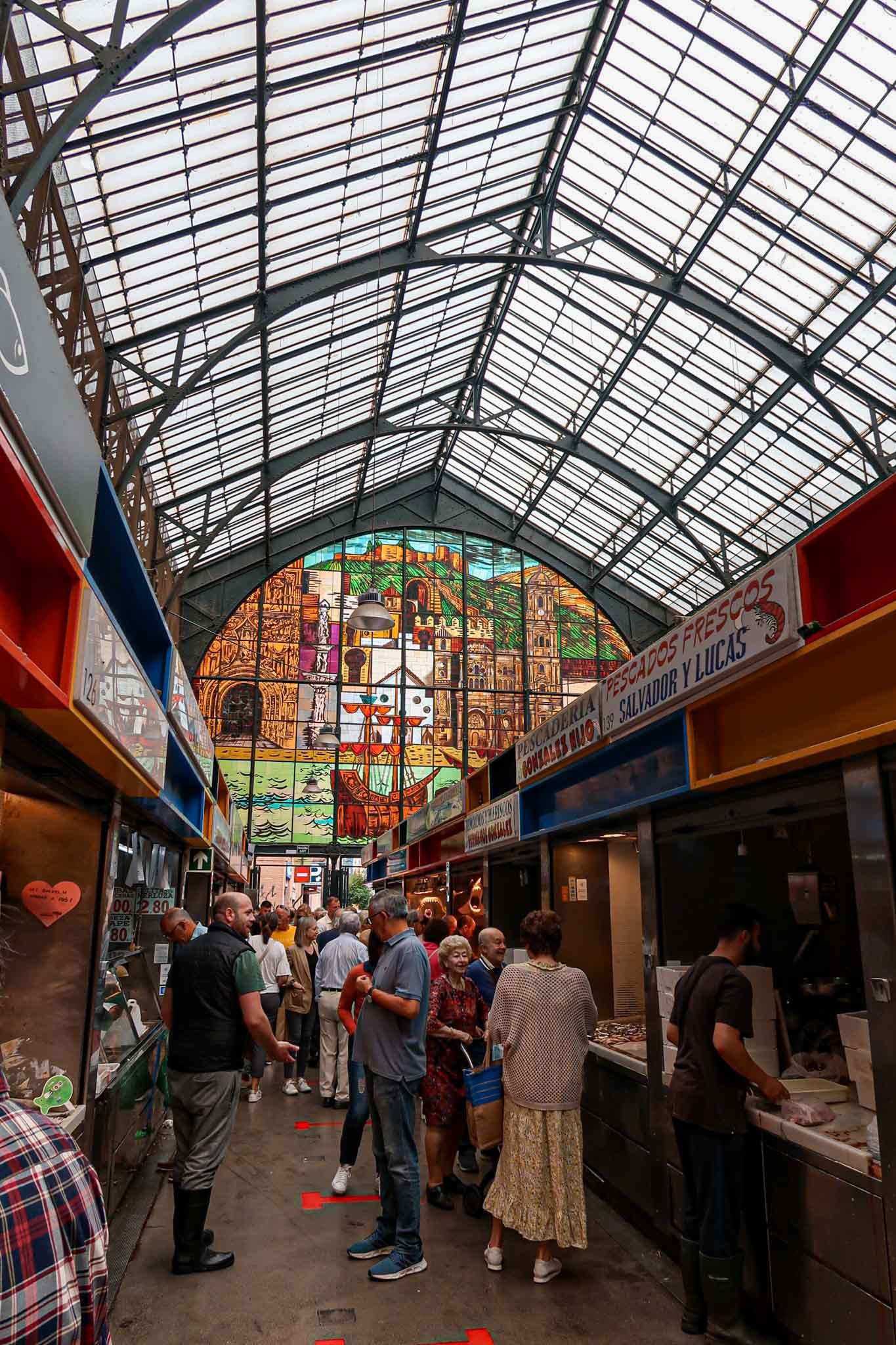 Inside Mercado de Atarazanas in Málaga, Spain - Yati Travel A bustling market scene inside Mercado de Atarazanas in Málaga, Spain, with a large crowd of shoppers and vendors. Best Things to Do in Málaga.