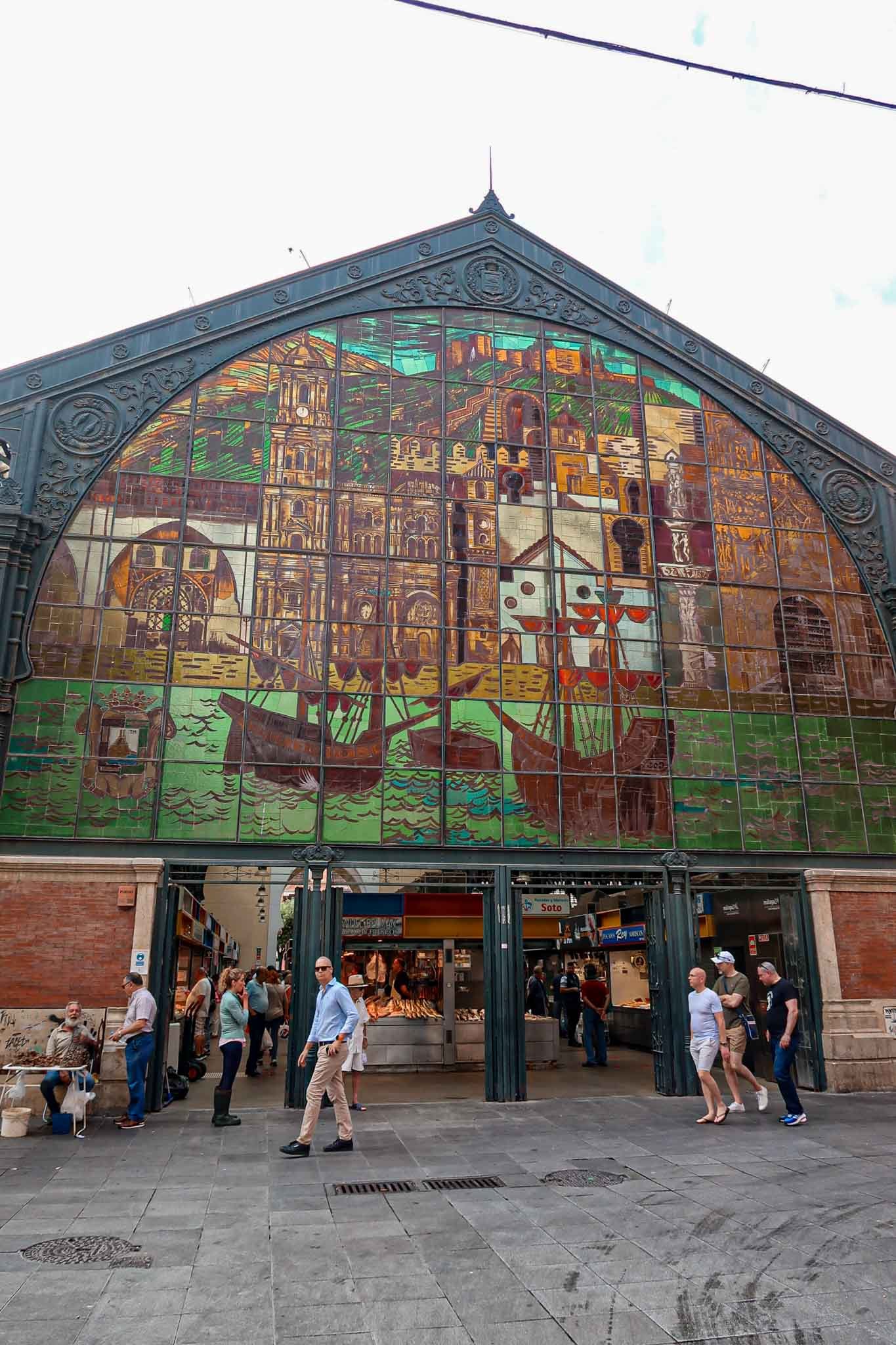Outside Mercado de Atarazanas in Málaga, Spain - Yati Travel The exterior of Mercado de Atarazanas in Málaga, Spain, showcasing a large building adorned with a colorful mural. Best Things to Do in Málaga.