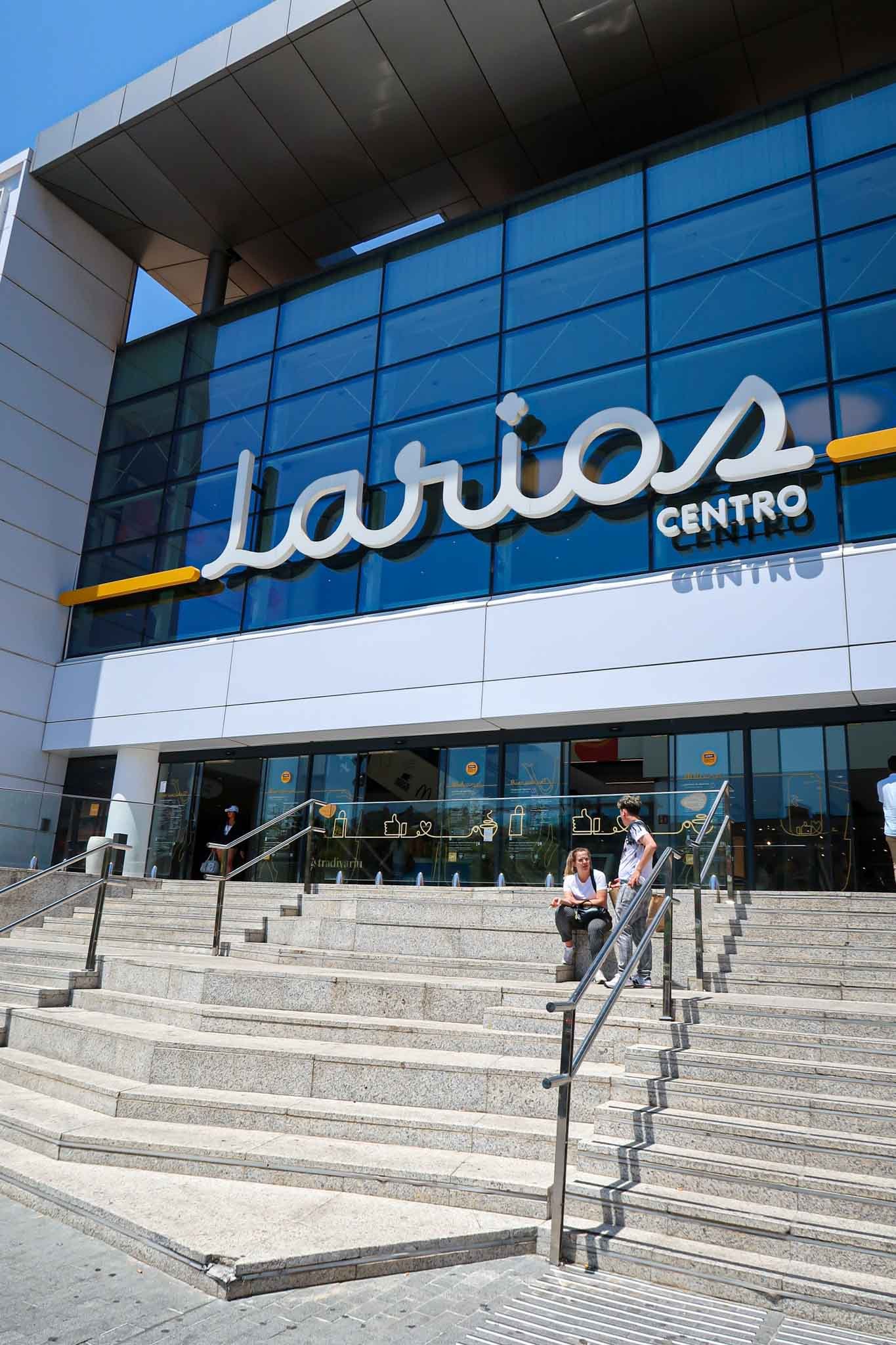Outside of Centro Comercial Larios Centro in Málaga, Spain - Yati Travel A man and woman sit on the steps outside Centro Comercial Larios Centro in Málaga, Spain, enjoying the sunny day. Best Things to Do in Málaga.