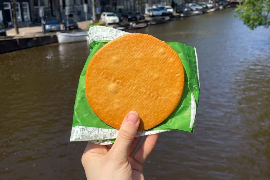 A person holds a traditional honey syrup waffle in front of a scenic canal in Amsterdam. Best Bakeries in Amsterdam.