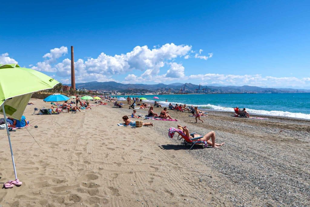 View of Playa la Malagueta in Málaga, Spain - Yati Travel A sunny view of Playa la Malagueta in Málaga, Spain, with people relaxing and sunbathing on the sandy beach. Best Things to Do in Málaga.